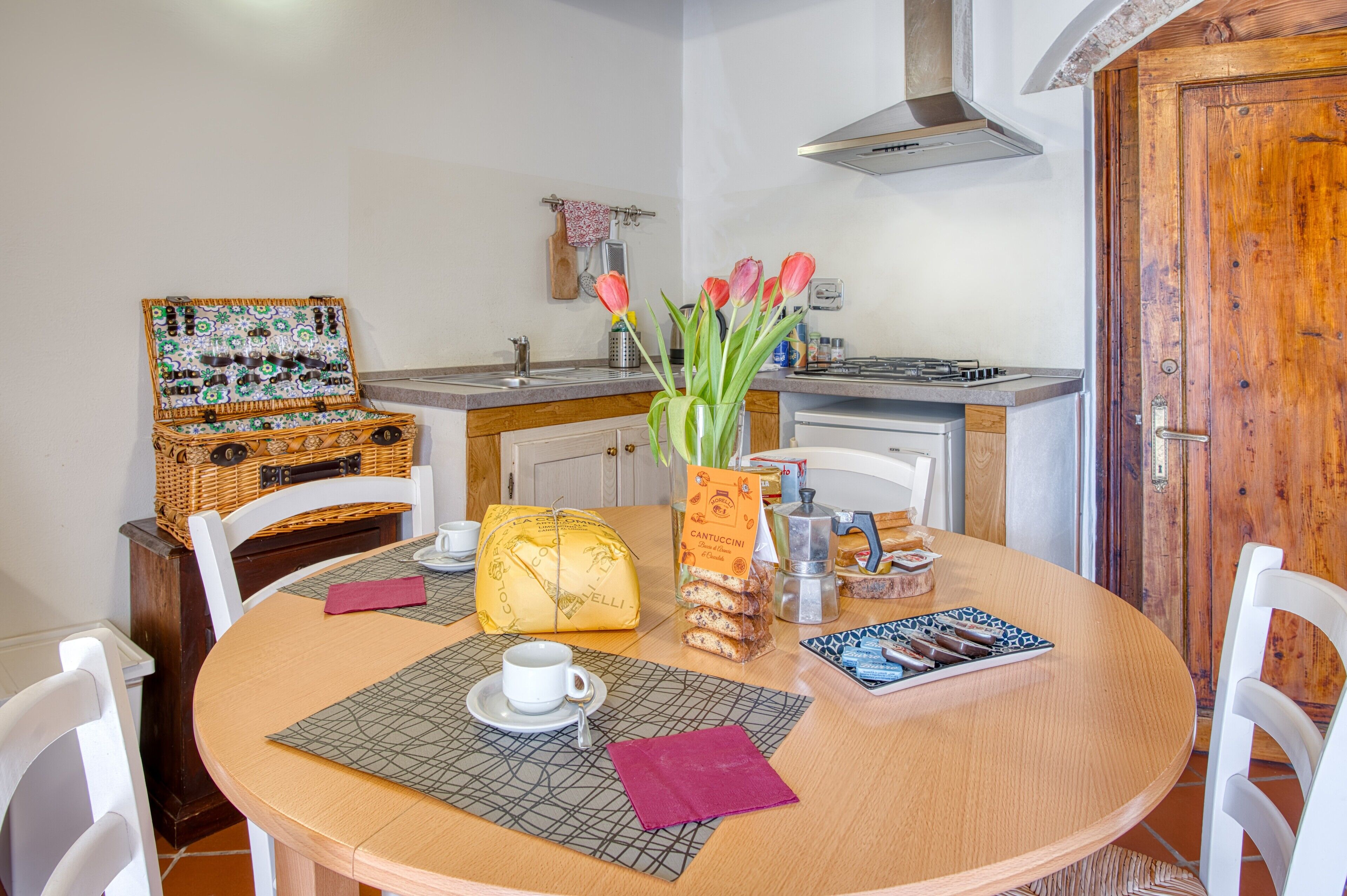 Een eetgedeelte met een ronde tafel, stoelen, een vaas met bloemen, een mand en een koffiezetapparaat.