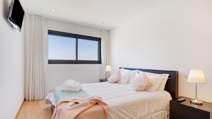 Une chambre d’hôtel avec un lit, un téléviseur fixé au mur, une fenêtre avec des rideaux et une table de chevet avec une lampe.