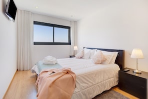 A hotel room with a bed, a TV mounted on the wall, a window with curtains, and a nightstand with a lamp. - Holiday Apartment 'Sublime Mundo' with Shared Pool, Balcony & Wi-Fi (Apúlia)