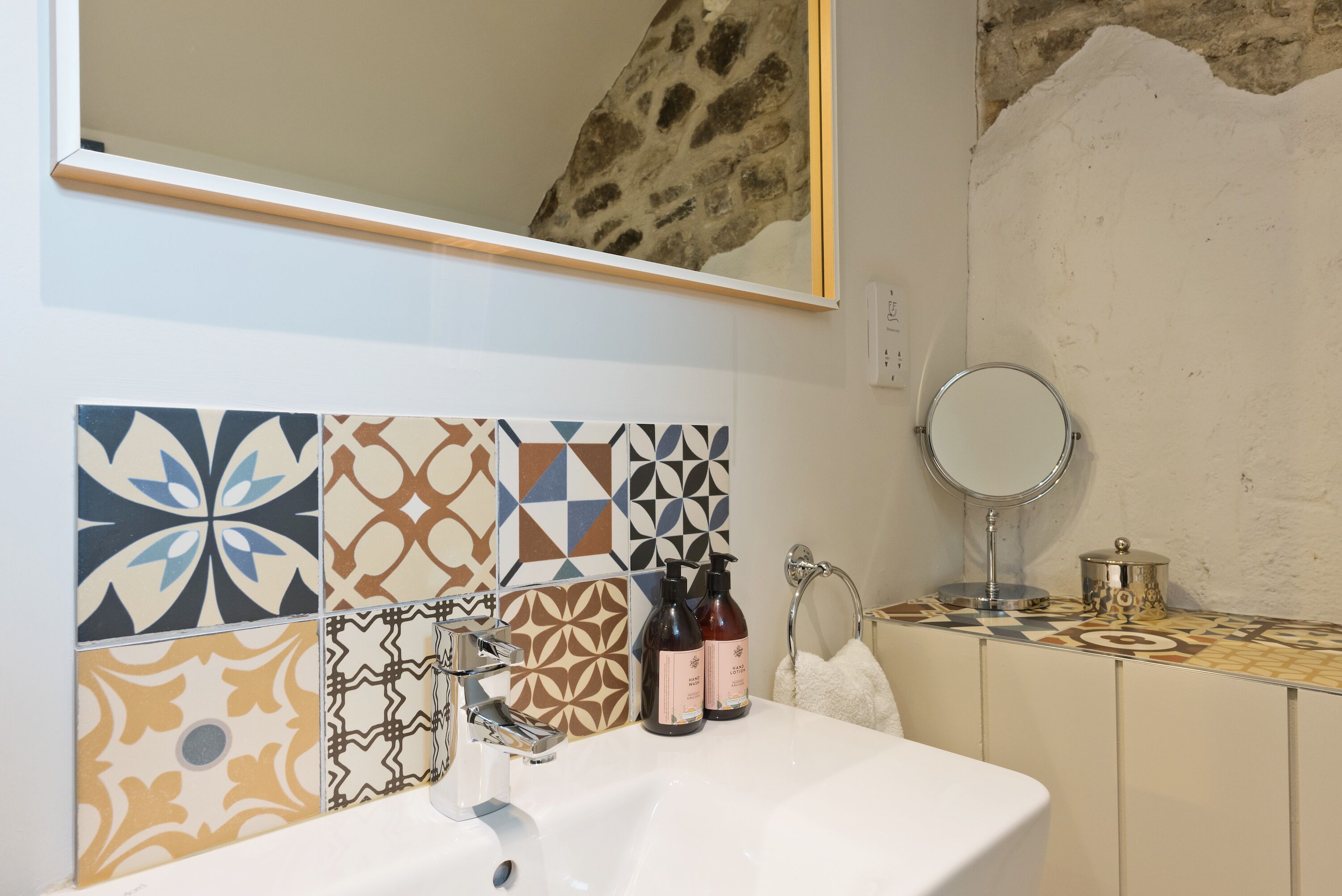 A bathroom with a white sink, patterned tiles, a mirror, and toiletries.