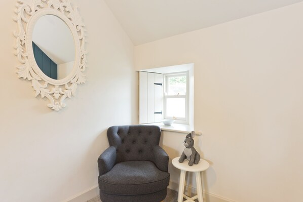 Una habitación de esquina con un sillón gris, un taburete blanco pequeño y un espejo blanco con detalles decorativos en la pared.