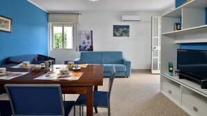 Una habitación con una mesa de comedor lista para dos personas, un sofá azul, un televisor sobre un mueble bajo y una ventana con vistas a la vegetación.