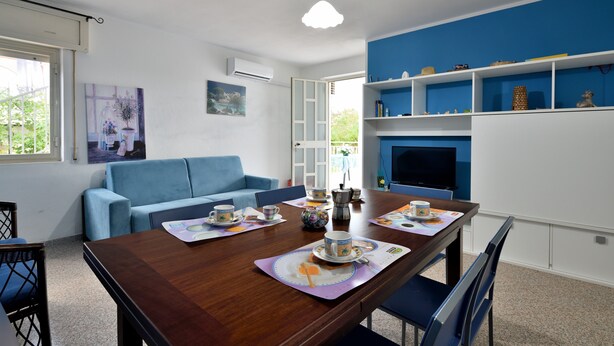 Un comedor con una mesa de madera dispuesta para cuatro personas, un sofá azul y un televisor sobre una pared azul.