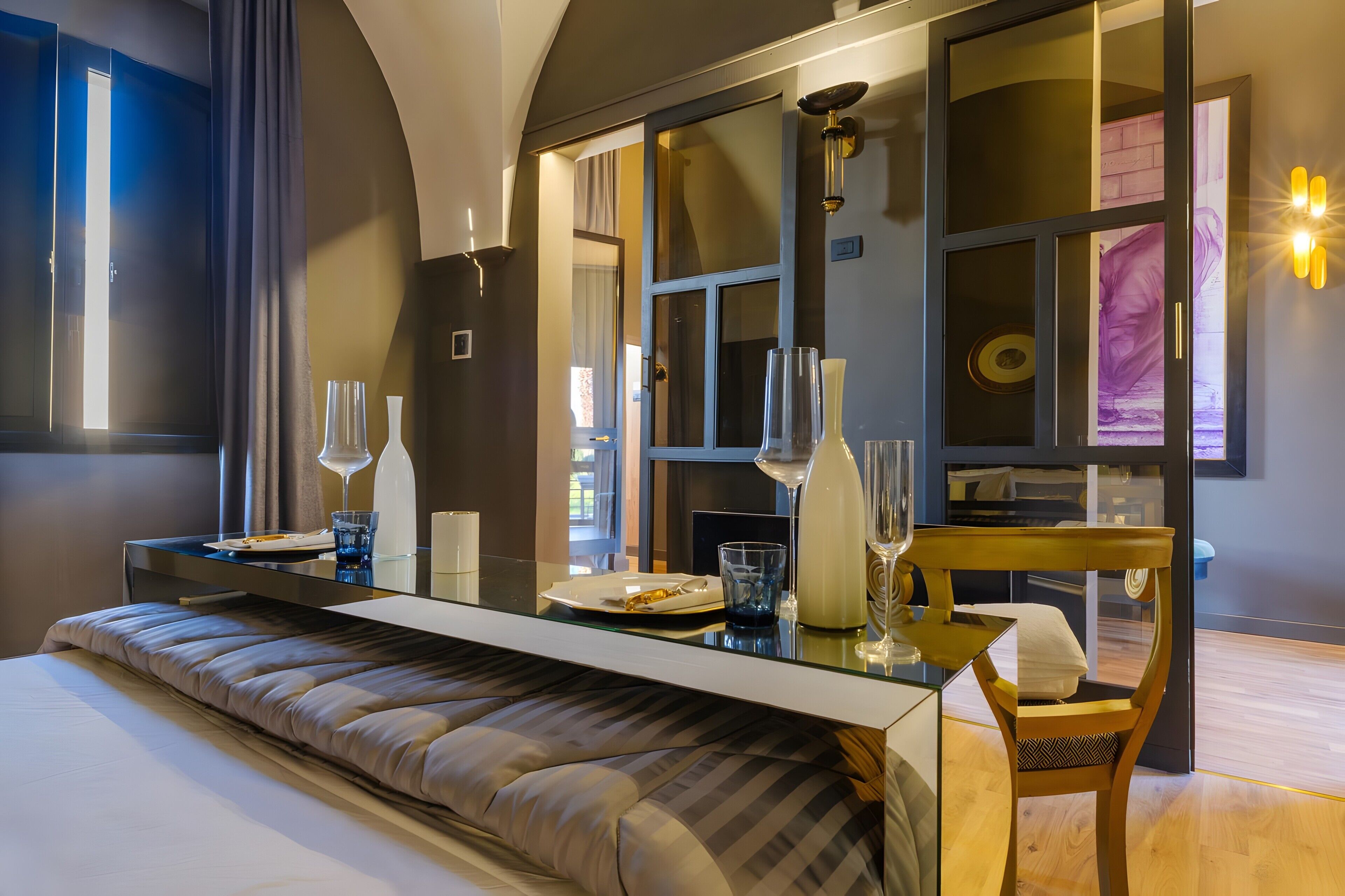 A modern hotel room with a large bed, a glass-top dining table, a seating area, and a view of the city.