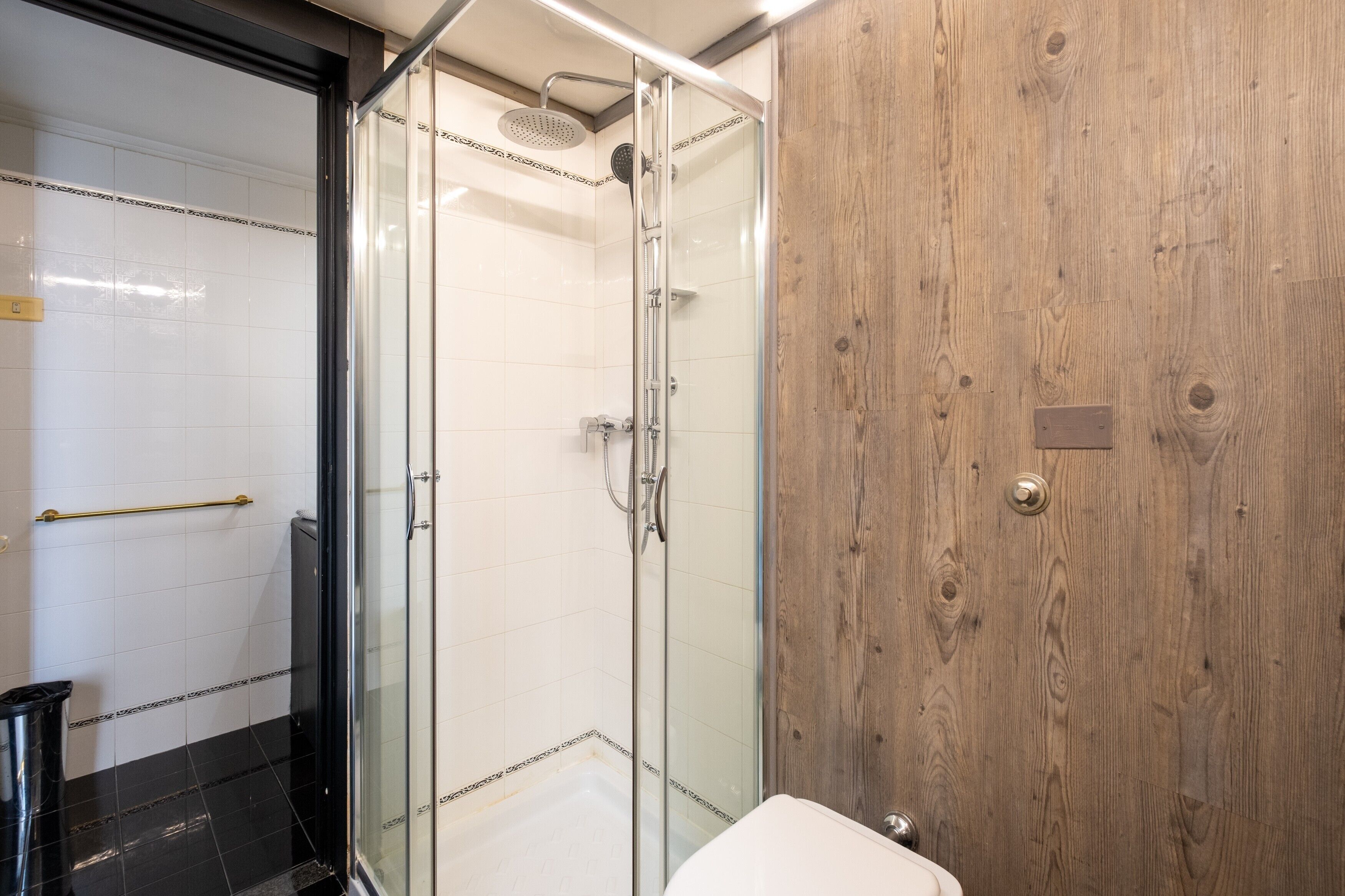 Eine begehbare Dusche mit Glaswänden, einer Handbrause, einer Holzwand und einem gefliesten Boden in Weiß.