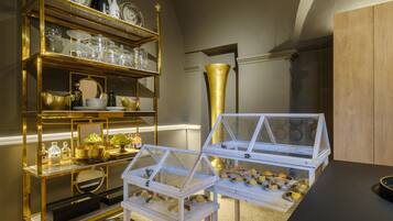 A room with a gold-framed display shelf, a glass display case with pastries, and a large golden vase.