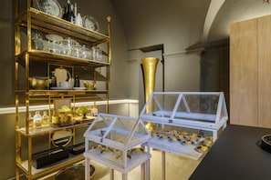 Une pièce comprenant une étagère décorative encadrée d’or, une vitrine en verre où sont exposées des pâtisseries, et un grand vase doré.