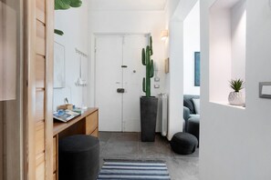 Un couloir avec un bureau en bois, un pouf noir, un tapis à rayures, un grand cactus et une porte blanche.