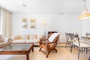 A living room with a sofa, a wooden chair, a glass coffee table, a dining table with chairs, and wall-mounted artwork.