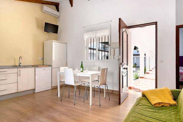 Une cuisine moderne avec des appareils électroménagers blancs, un coin repas avec une table et des chaises, et un canapé vert agrémenté d’un plaid jaune.