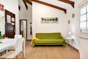 A room with a green sofa, a small table with a lamp, a white dining table with chairs, and a wall-mounted plant display.