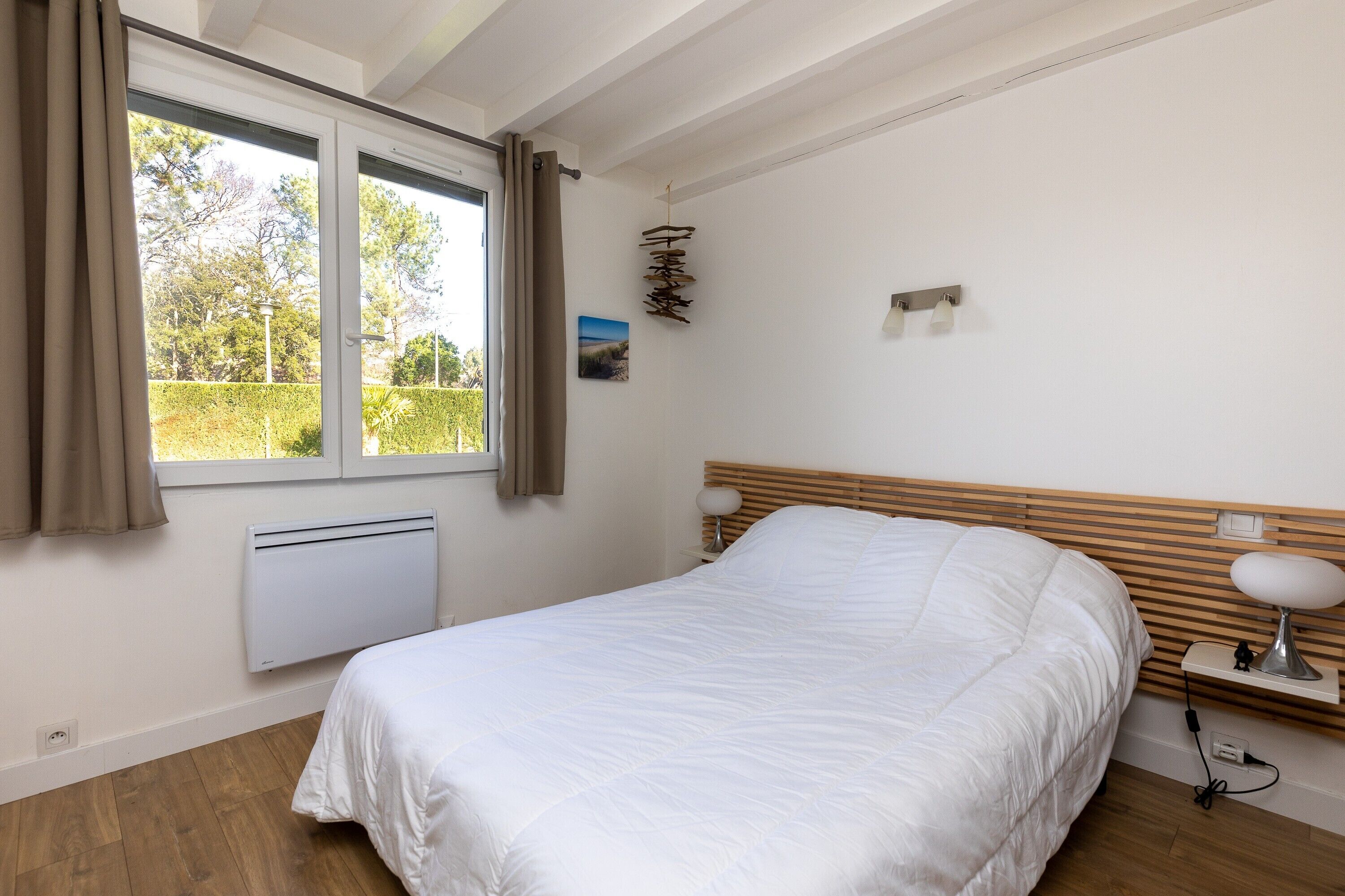 A bedroom with a bed, a window with curtains, a radiator, a bedside table with a lamp, and a wall-mounted light fixture.