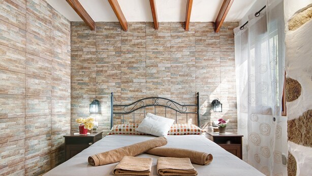 Um quarto com parede de tijolos, uma cama com toalhas, uma mesa de cabeceira com abajur e uma janela com cortina.