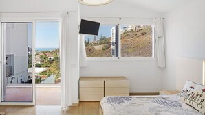 Camera da letto con un letto, una TV, un comodino, una cassettiera e una finestra che si affaccia su un pendio e degli edifici.