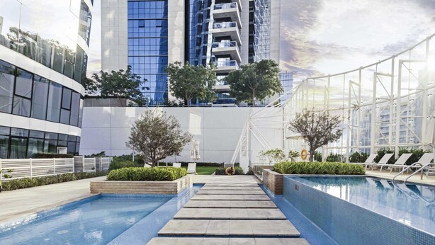 A modern outdoor swimming pool area with a clear sky, surrounded by high-rise buildings and lounge chairs. - LUX - The Pad Executive Suite 4 (Dubai)