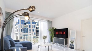 A modern living room with a sofa, a floor lamp, a TV, and a large window with a view of a building. - LUX - The Pad Executive Suite 4 (Dubai)