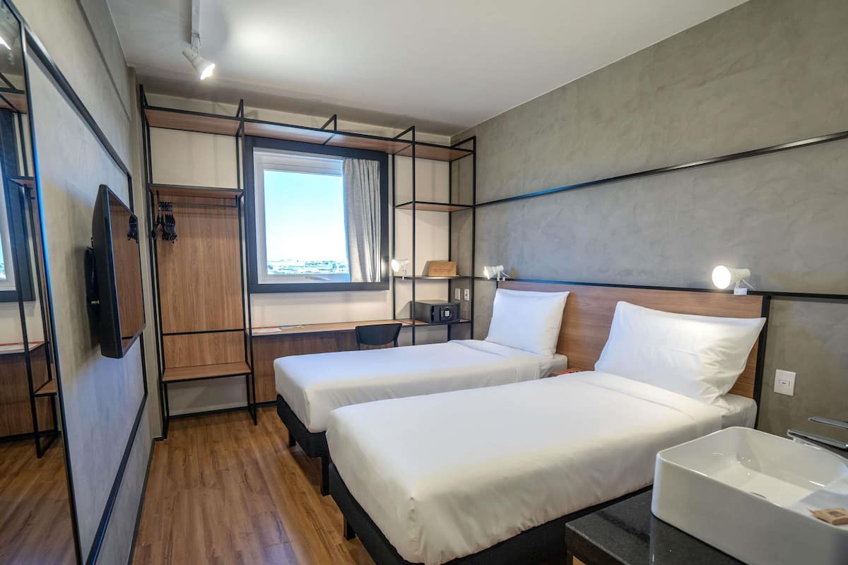 A modern hotel room with two single beds, a desk, a TV, a window with a view, and a bathroom with a sink.