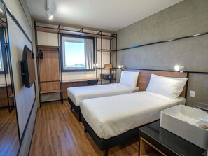 A modern hotel room with two single beds, a desk, a TV, a window with a view, and a bathroom with a sink.