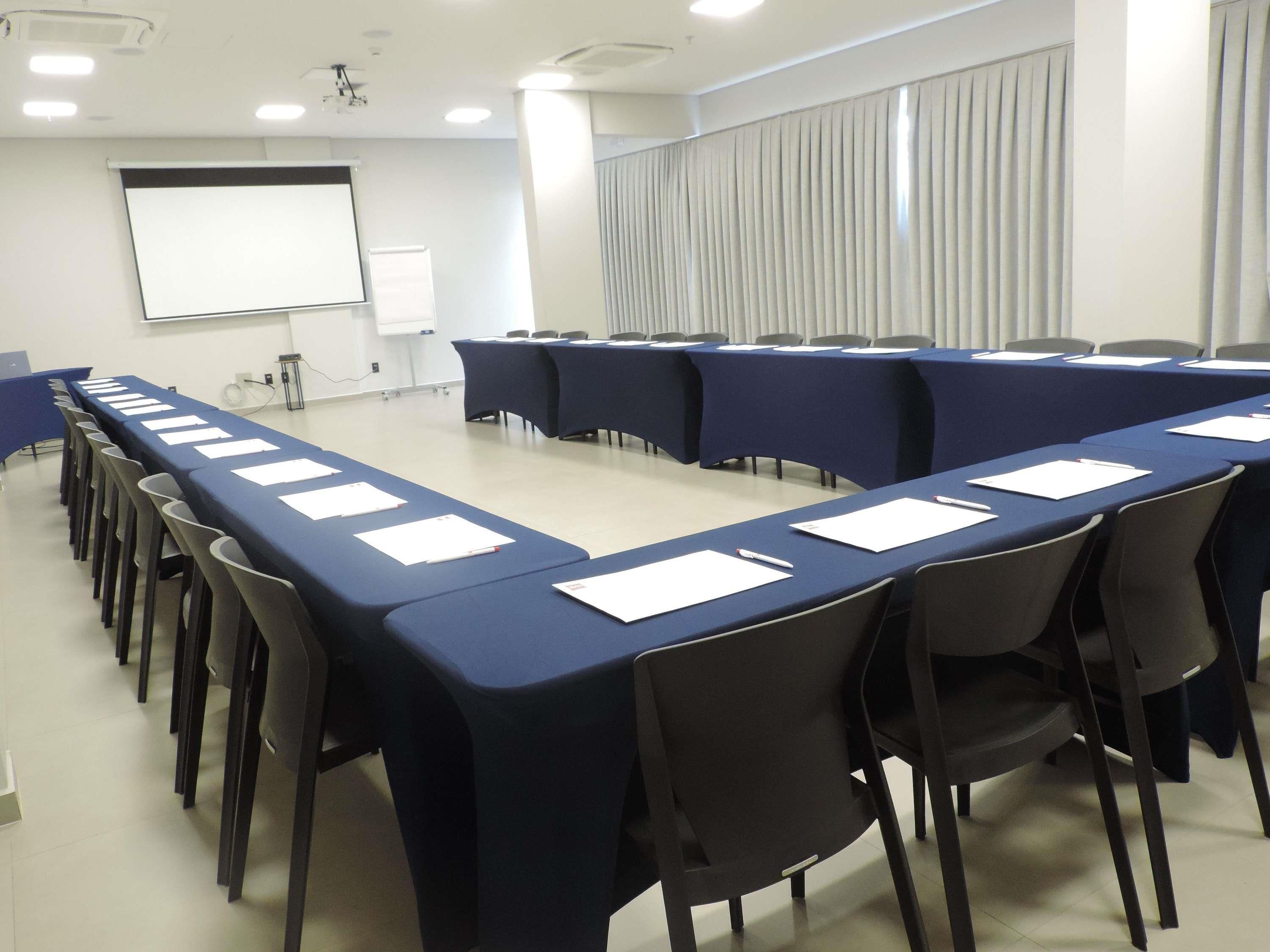 Una sala de conferències amb taules llargues, cadires, una pantalla de projector i una pissarra.