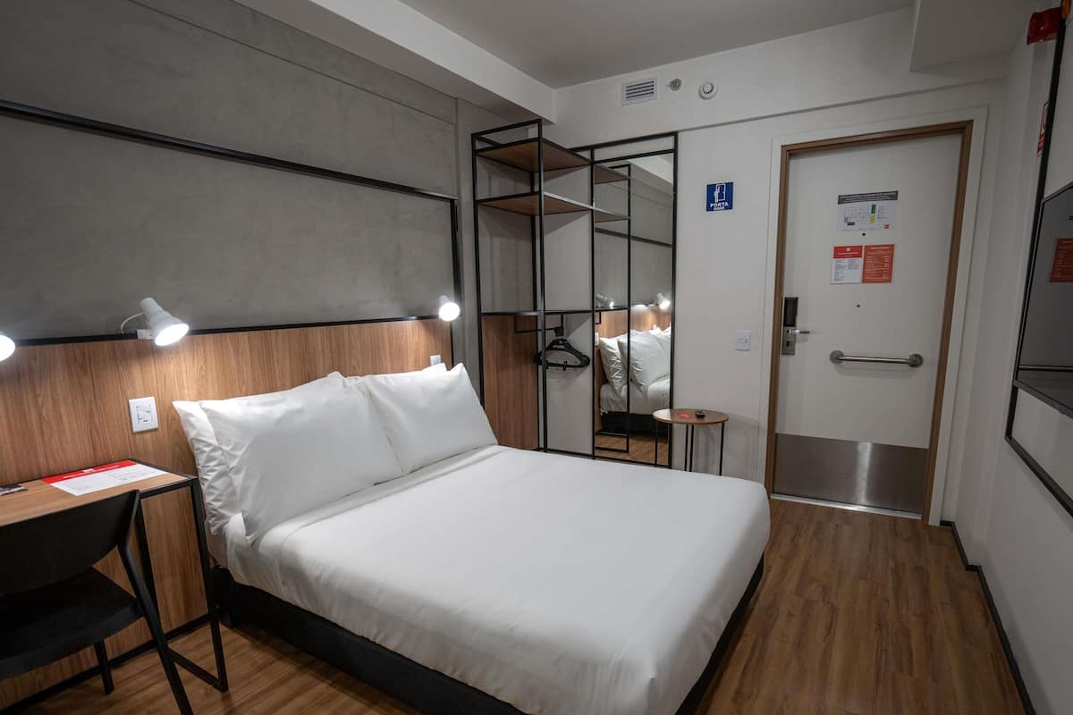 Quarto de hotel com uma cama, mesa de cabeceira e escrivaninha com uma cadeira. Há uma porta com puxador e uma pequena mesa com cadeira ao lado.