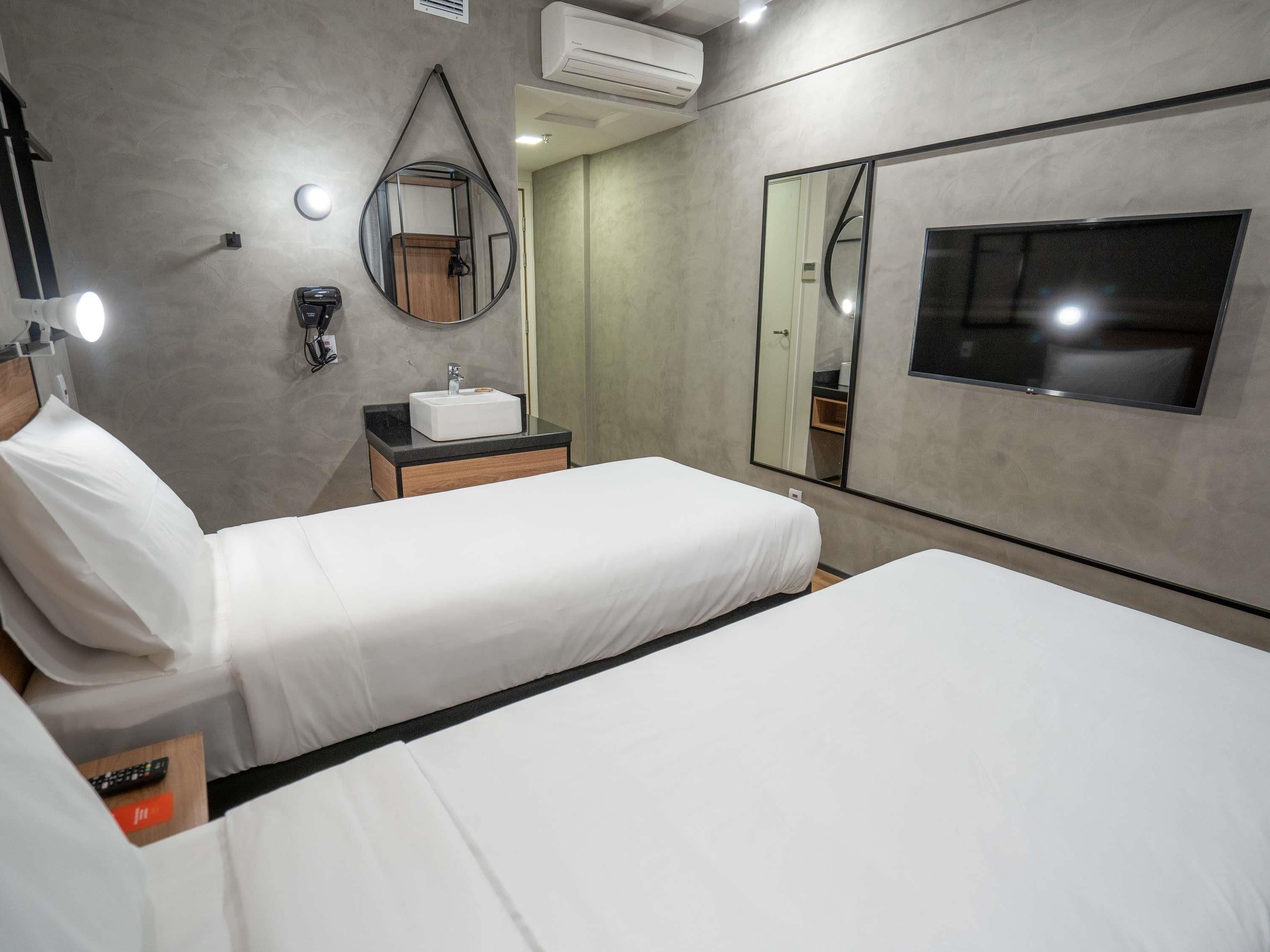 A hotel room with two beds, a bedside table with a white sink, a mirror, and a TV.