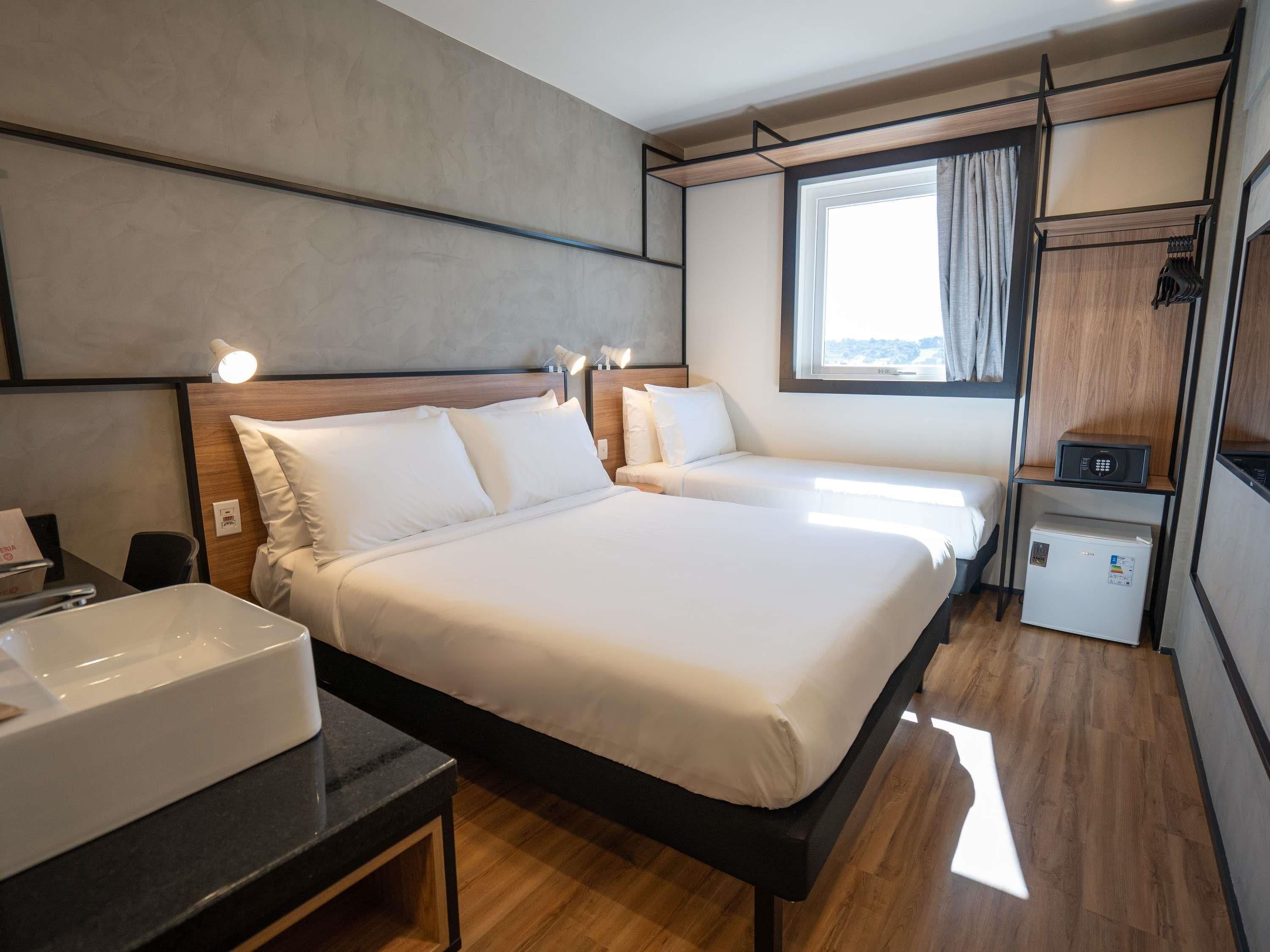 A modern hotel room with a large bed, bedside tables, a built-in heater, a small refrigerator, and a window with curtains.