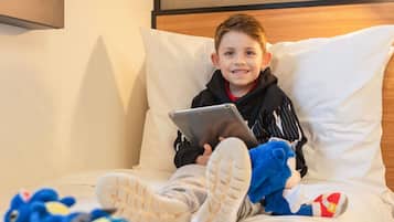 Un niño sentado en una cama con una tableta, usando sudadera con capucha y pantuflas.