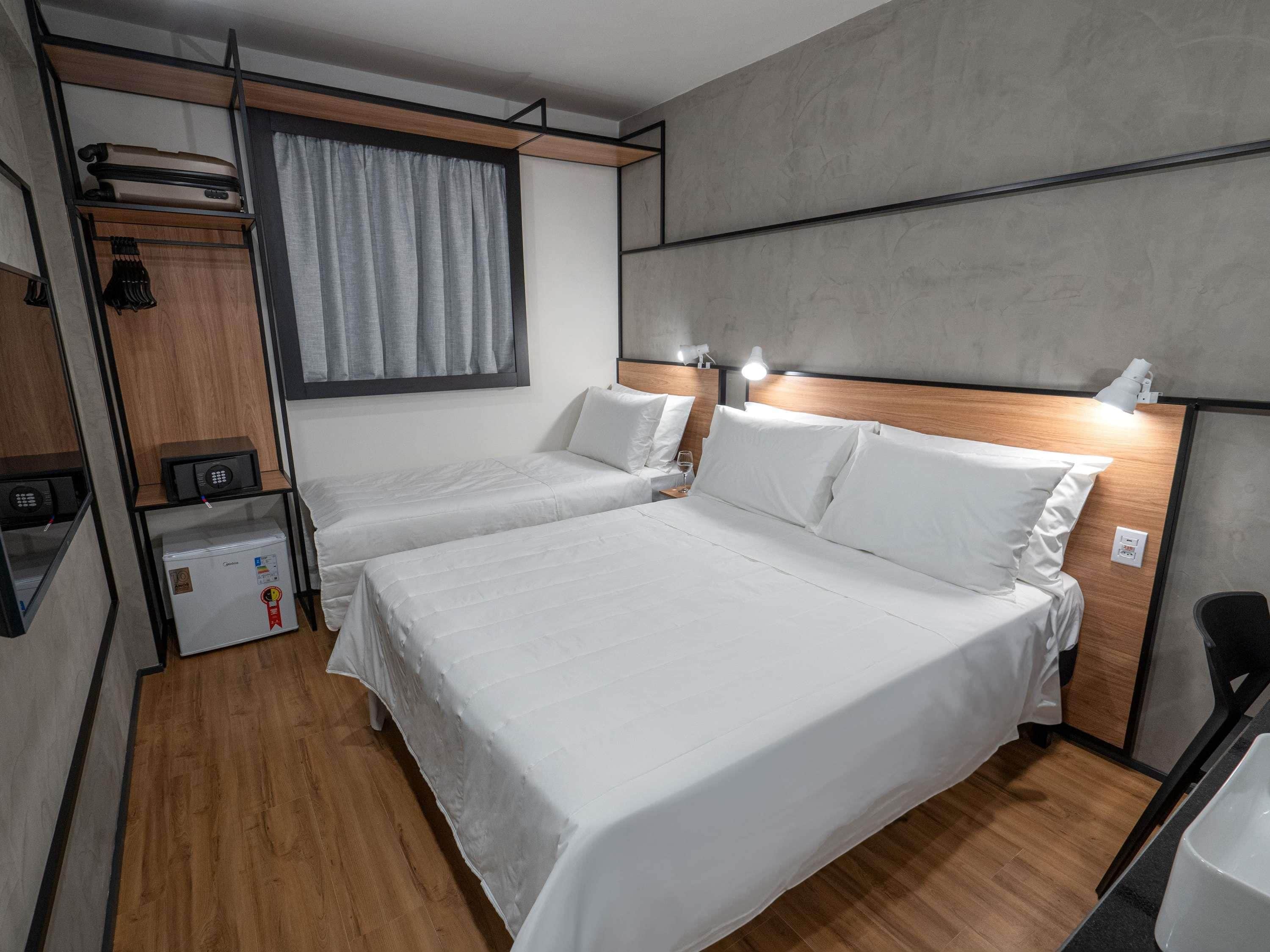 A modern hotel room with two beds, a small desk, a wardrobe, and a window with curtains.