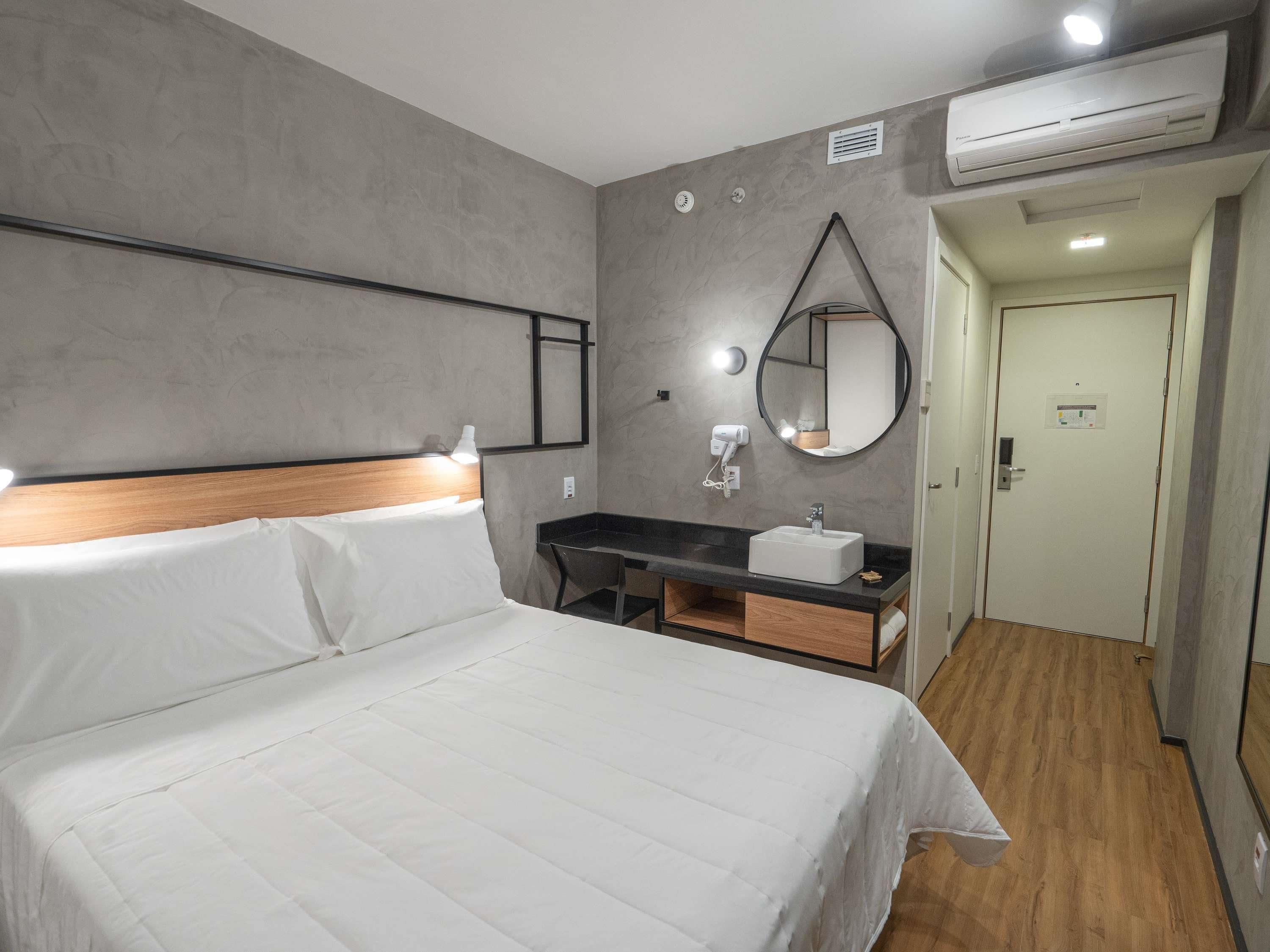 A hotel room with a bed, a desk with a sink, a mirror, and an air conditioning unit.