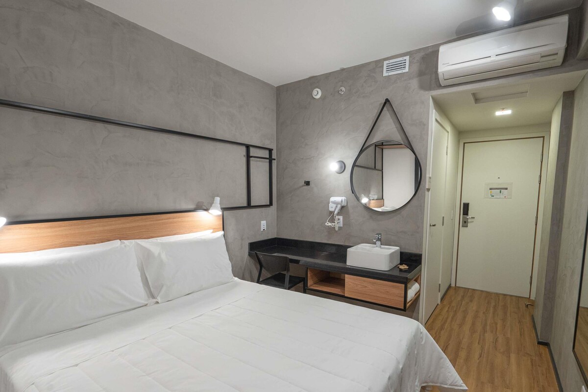 A hotel room with a bed, a desk with a sink, a mirror, and an air conditioning unit.