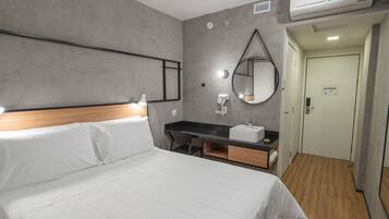 A hotel room with a bed, a desk with a sink, a mirror, and an air conditioning unit.