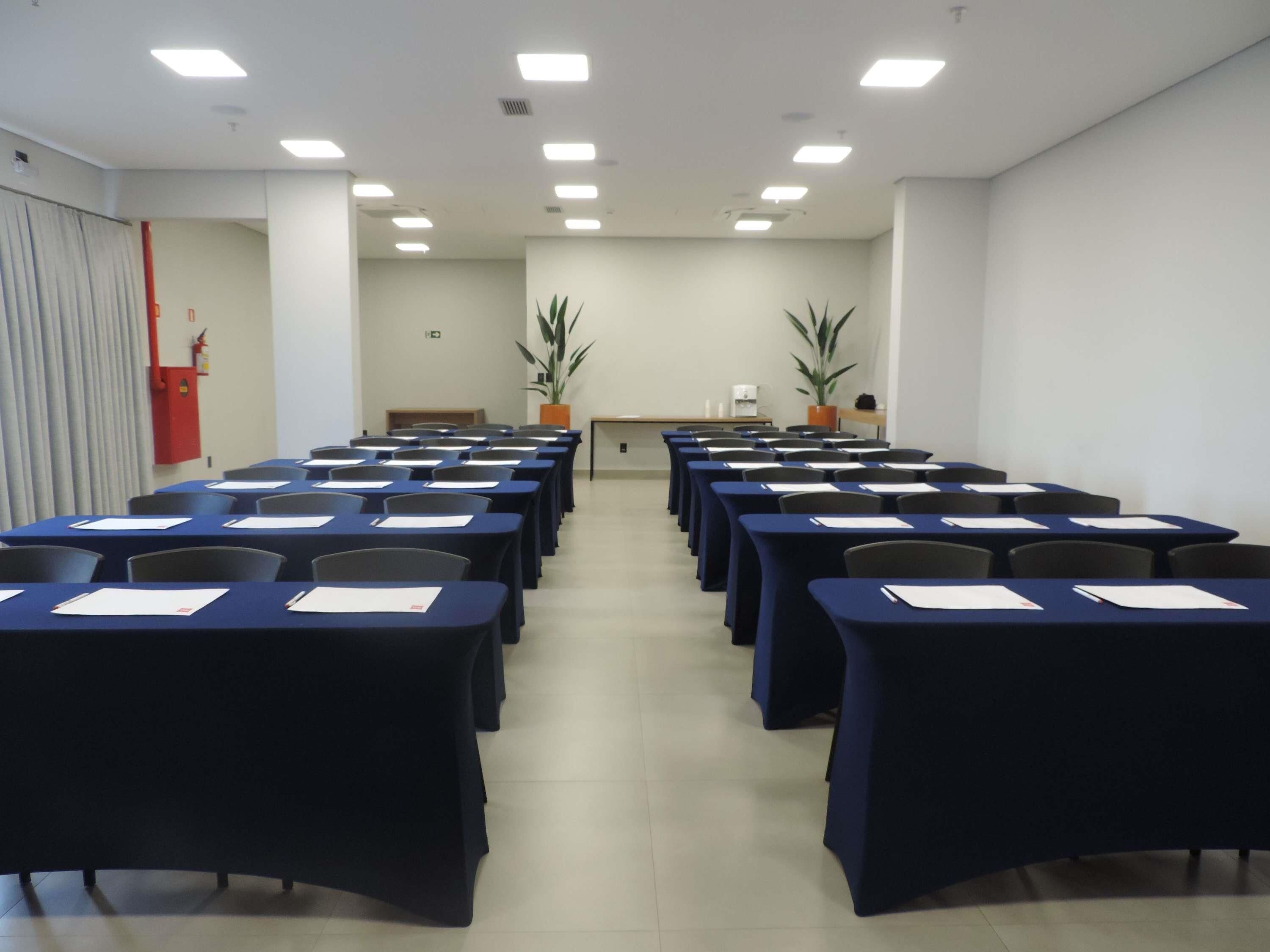 A conference room with rows of tables and chairs, a fire extinguisher, and two potted plants.