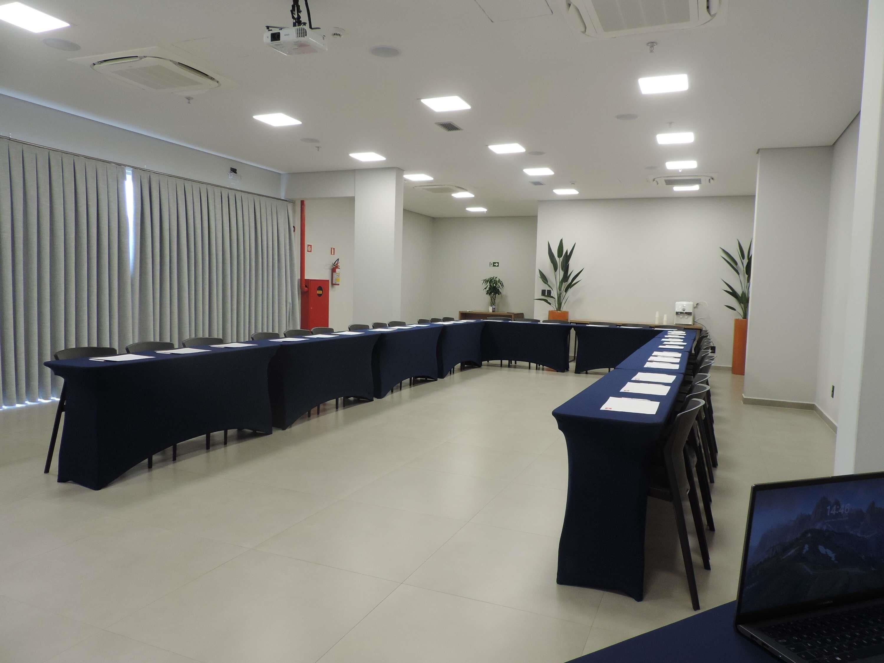 Uma sala de conferências com uma mesa comprida, cadeiras, um projetor e plantas em vasos.