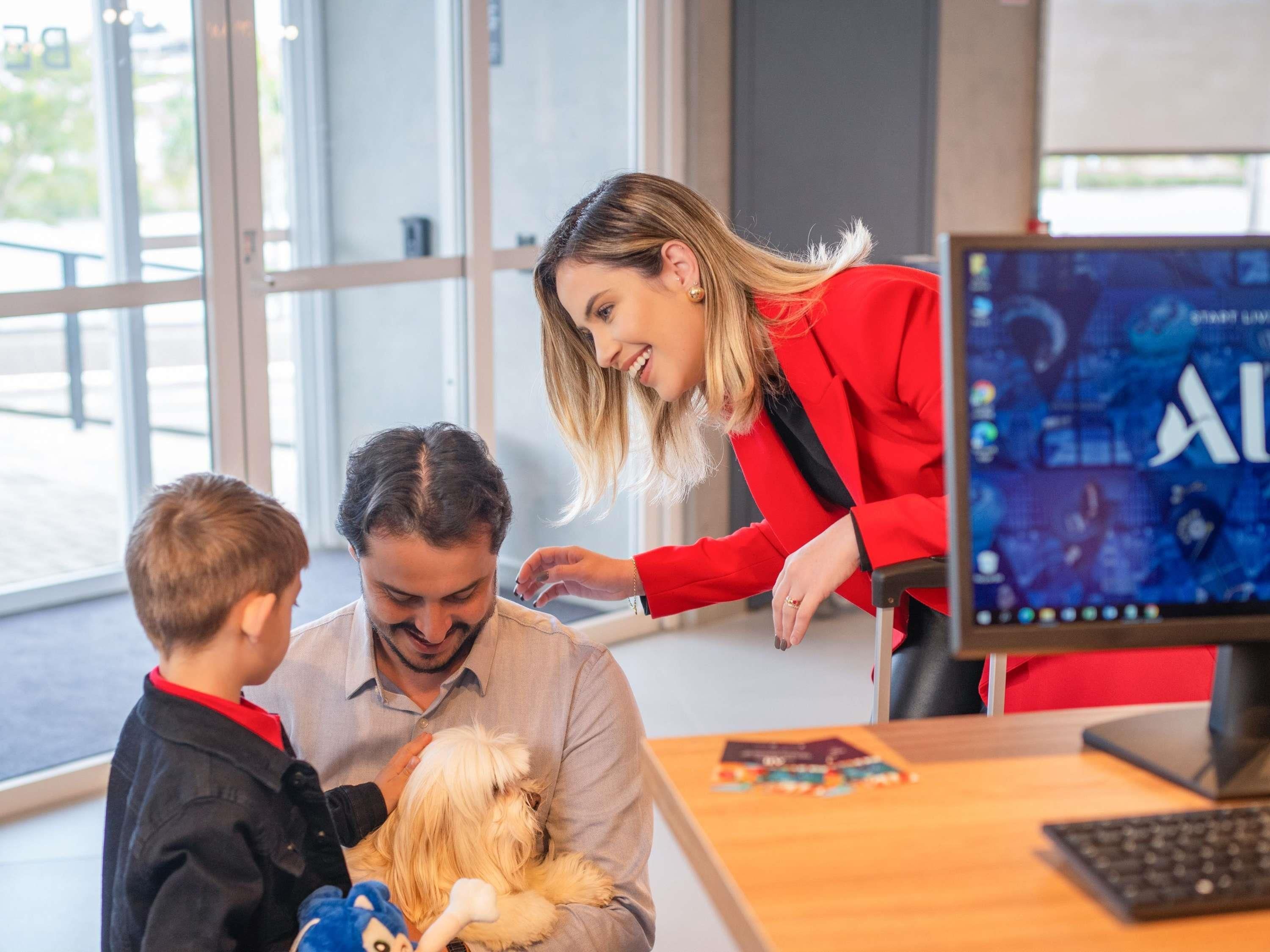 Una mujer con una chaqueta roja interactúa con un hombre y un niño, mientras un monitor muestra un videojuego al fondo.