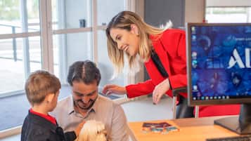 Una mujer con una chaqueta roja interactúa con un hombre y un niño, mientras un monitor muestra un videojuego al fondo.