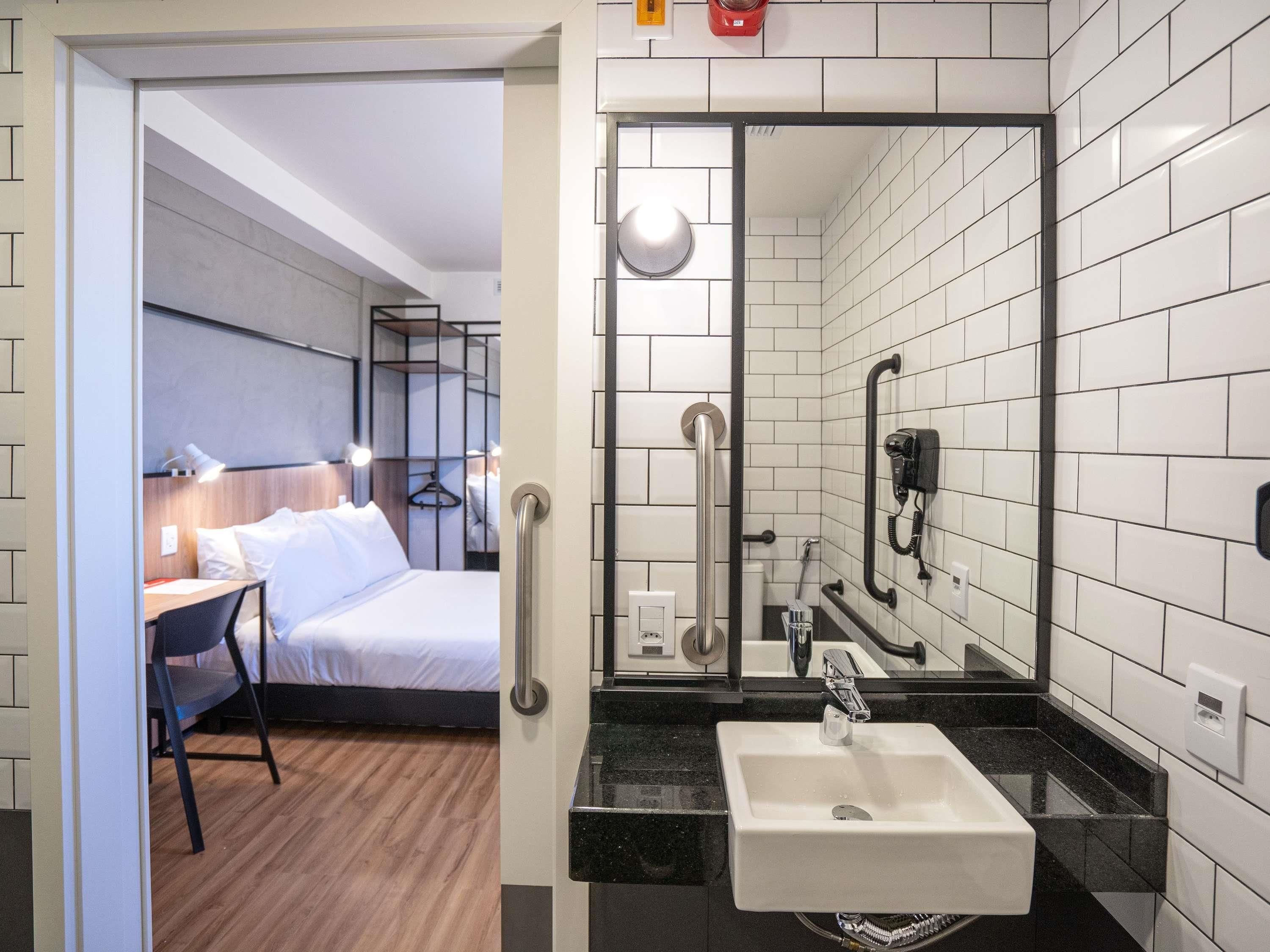 A hotel room with a bed, bedside table, lamp, and a bathroom with a sink, mirror, and towel rack.