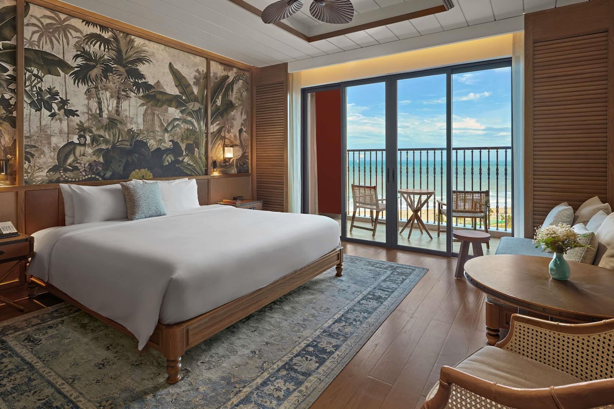 Habitación de hotel con una cama grande, un balcón con vistas a la playa, un área de comedor y un mural tropical en la pared.