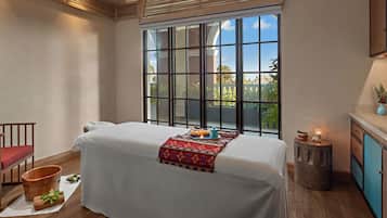 Relax in this serene spa room with a view of lush greenery and a clear sky, featuring a single bed, a wooden bench, and a decorative rug.