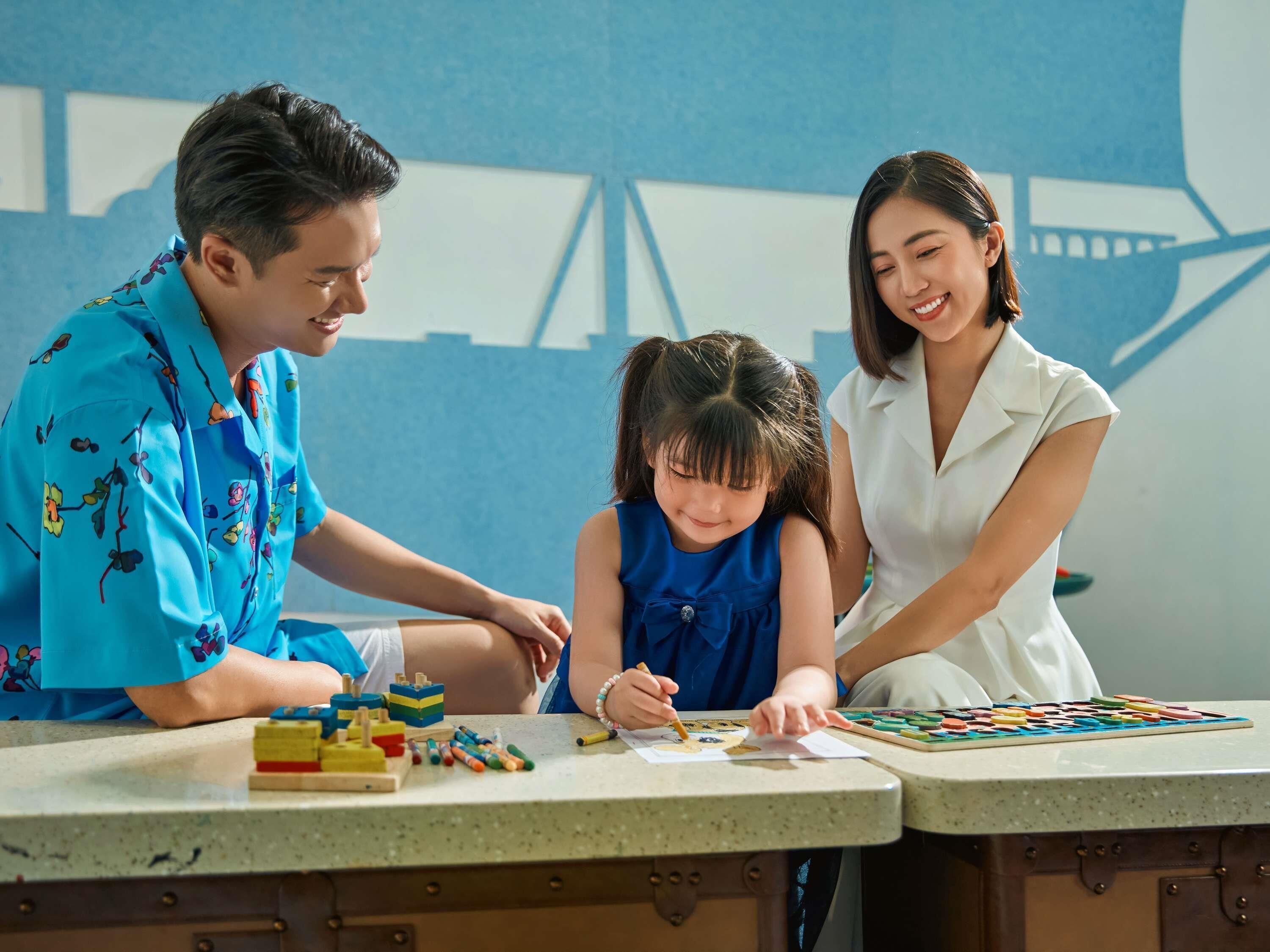 A family engaged in a board game at a table with educational toys and a cityscape mural in the background.