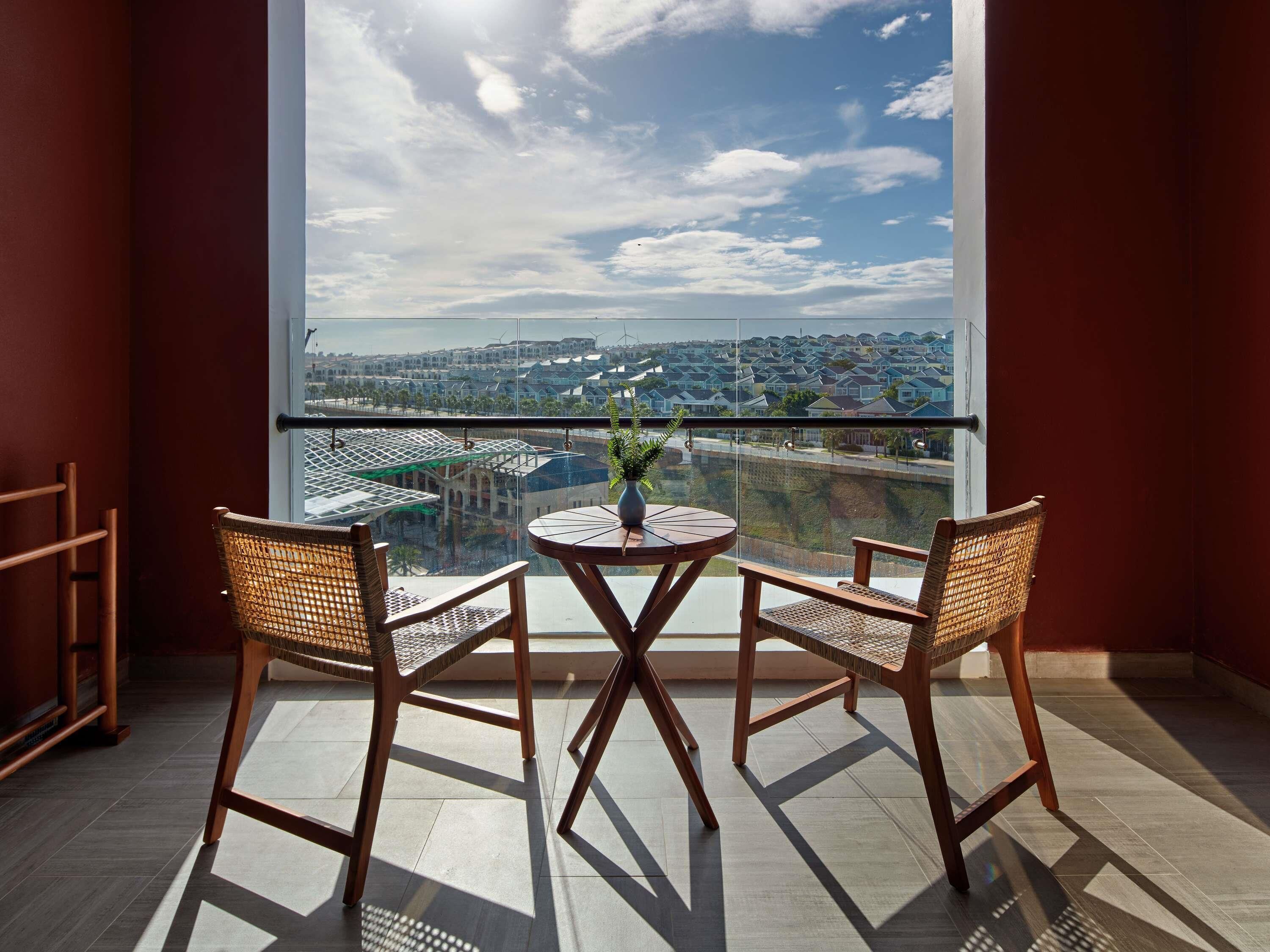 A balcony with two wicker chairs and a small round table, overlooking a residential area with houses and greenery.
