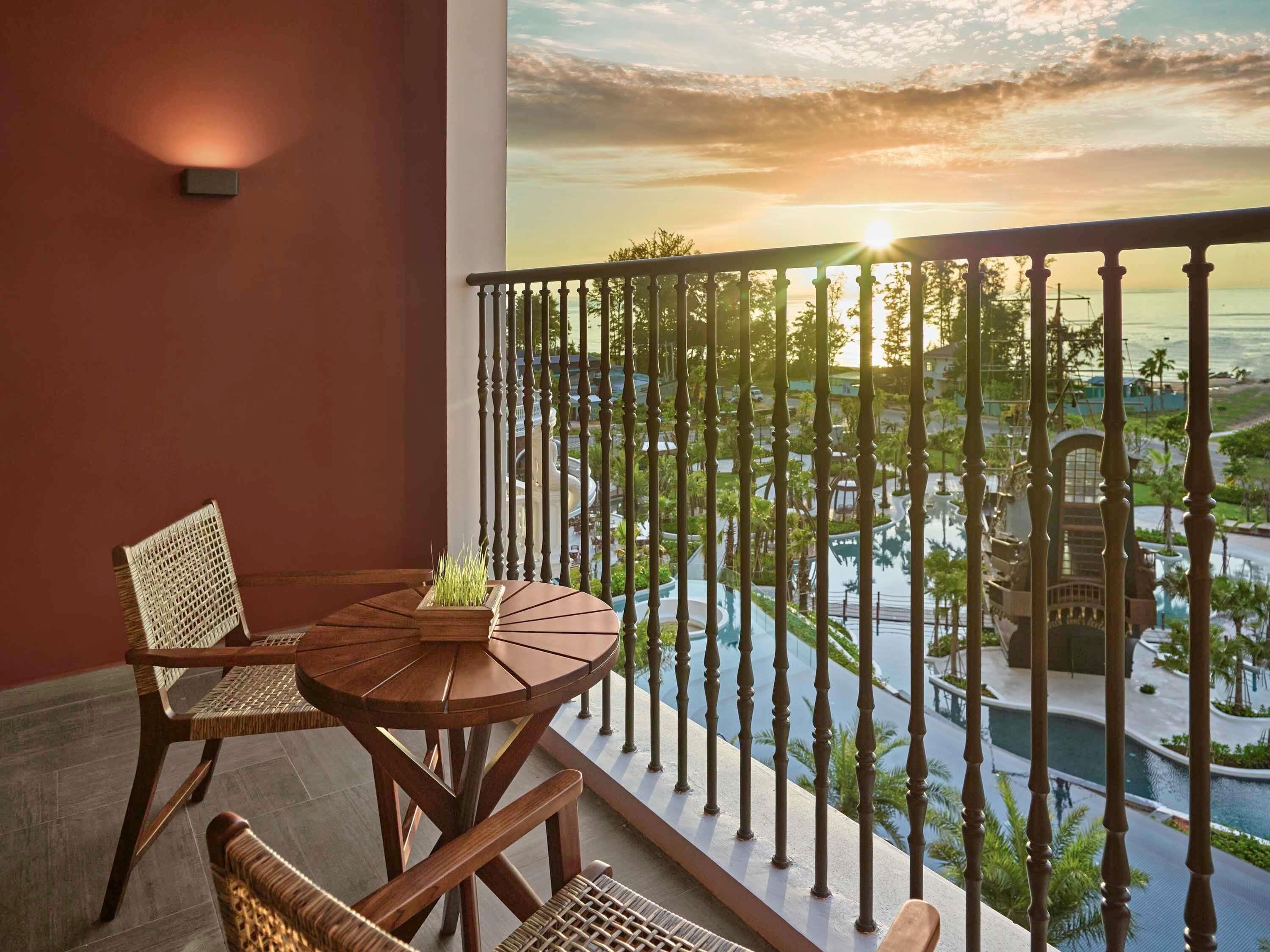 Een balkon met een ronde houten tafel en twee rieten stoelen, met uitzicht op een zwembad en het resort.