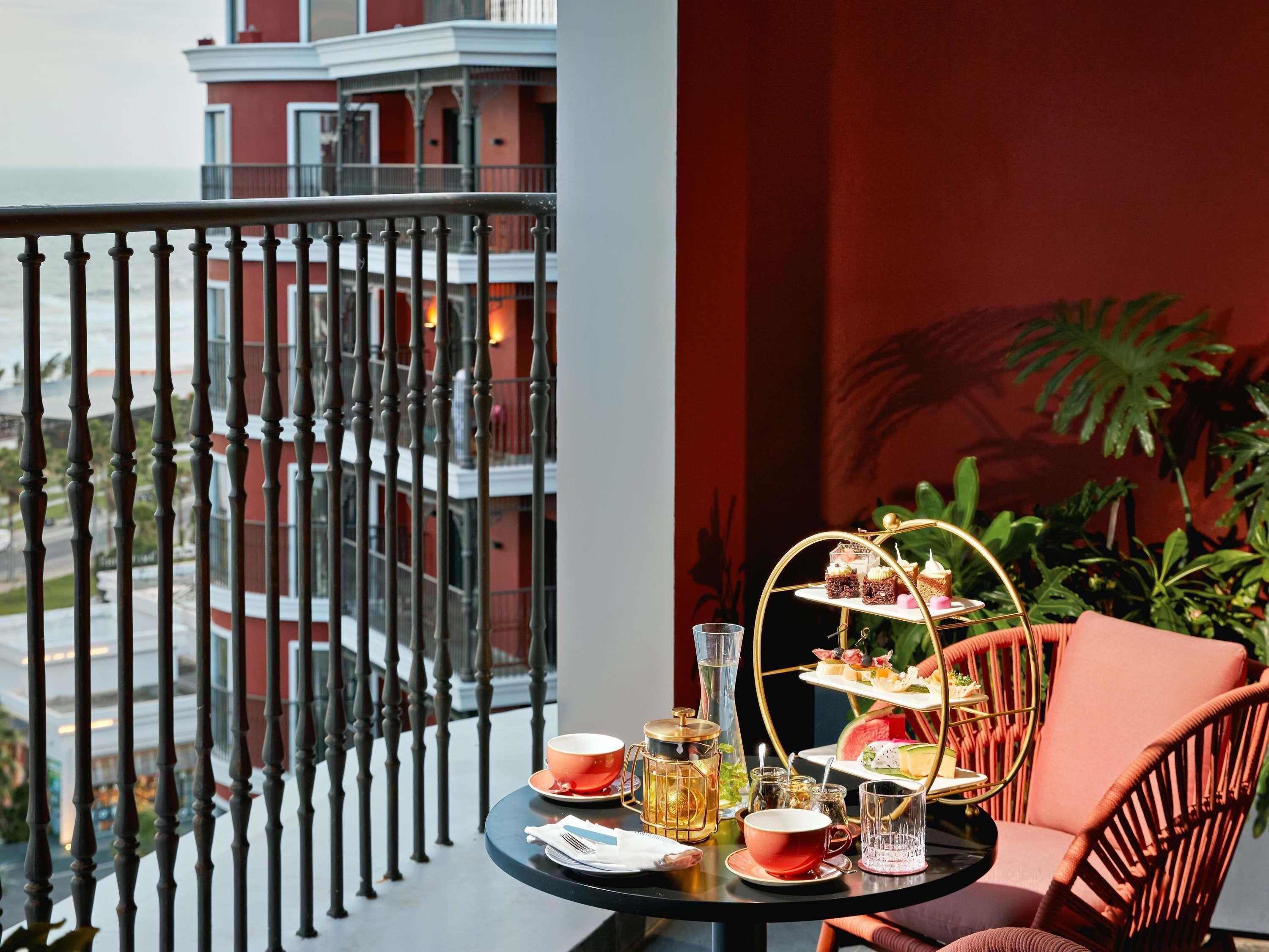 A balcony with a table set for tea, a three-tiered dessert stand, and a view of a building.