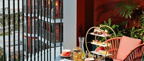 A balcony with a table set for tea, a three-tiered dessert stand, and a view of a building.