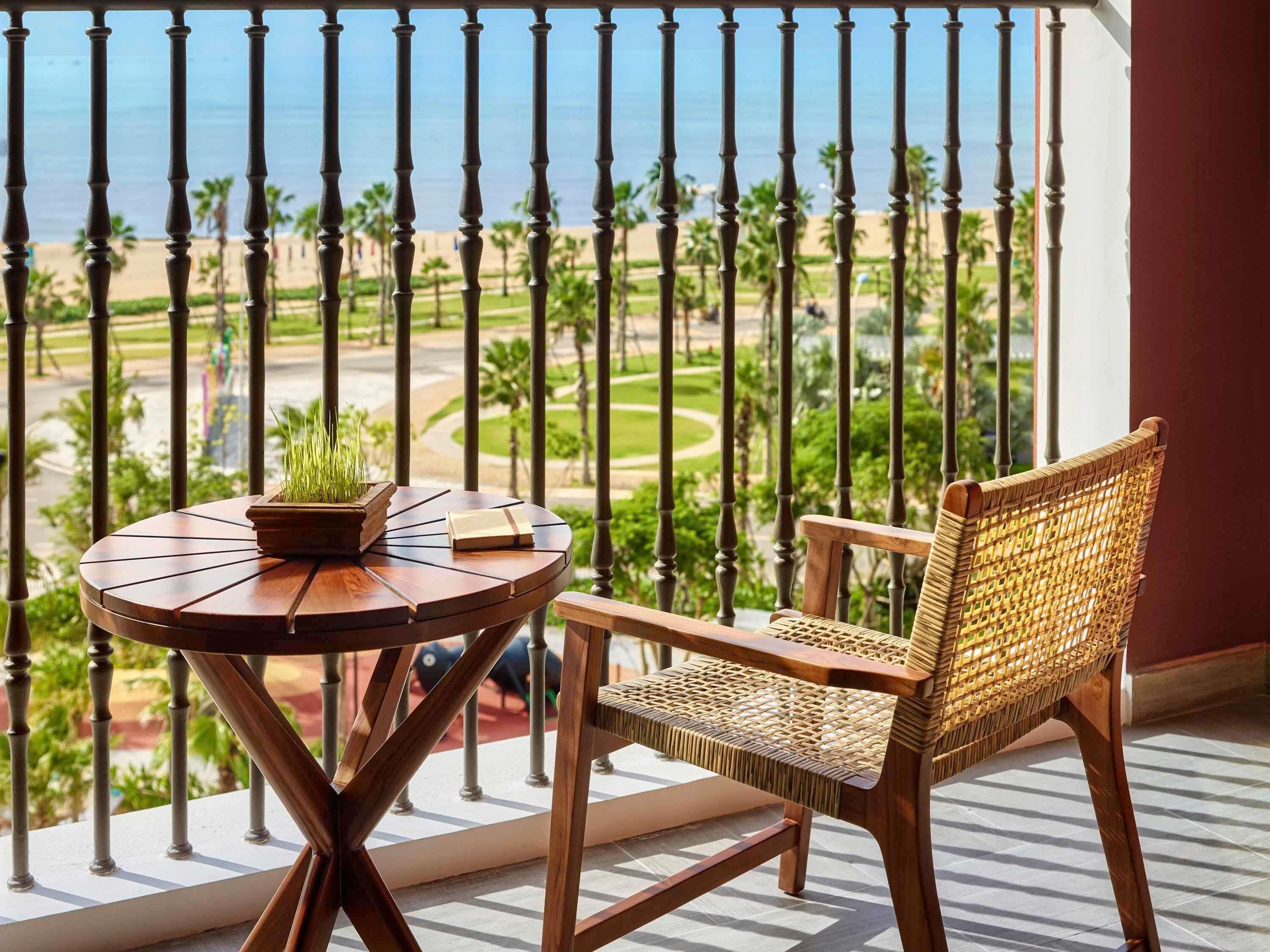 Un balcón con vista a la playa, una mesa redonda de madera con una planta y un libro, y una silla de mimbre.