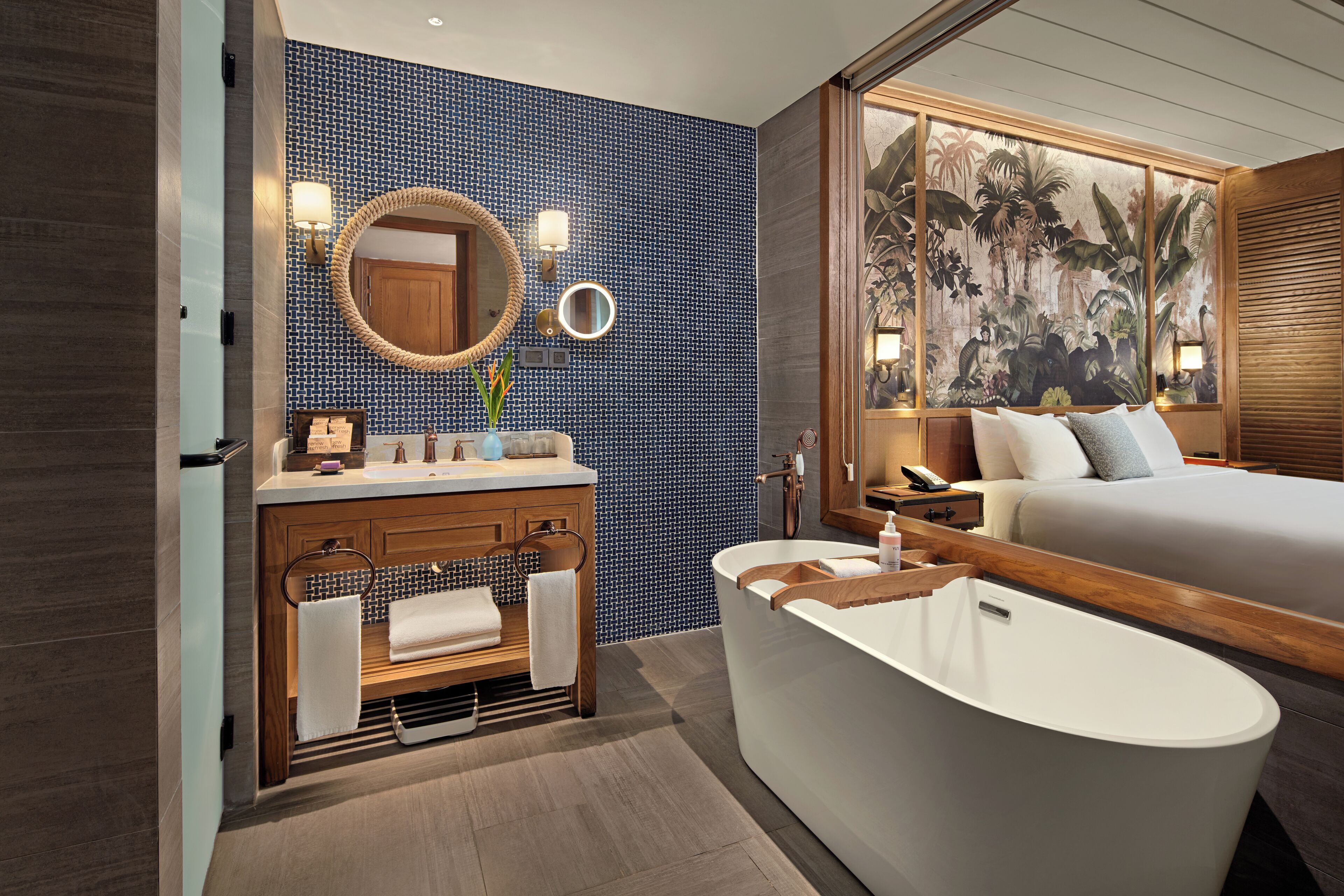Un baño moderno con una bañera independiente, un tocador de madera con espejo y una cama grande con un mural tropical.