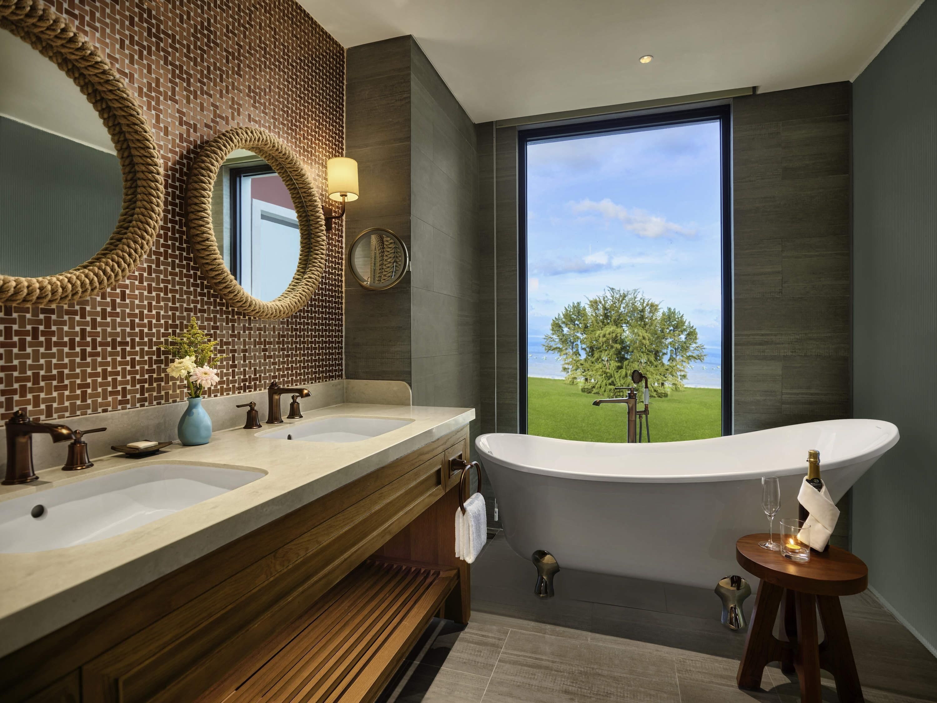 A modern bathroom with a large window, two sinks, a bathtub, and a wooden vanity.