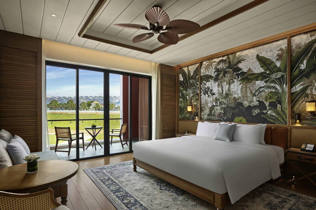 Habitación de hotel con una cama grande, un balcón con mesa y sillas, vistas a casas y un mural tropical.