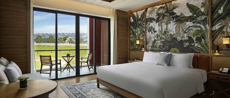 A hotel room with a large bed, a balcony with a table and chairs, a view of houses, and a tropical mural.