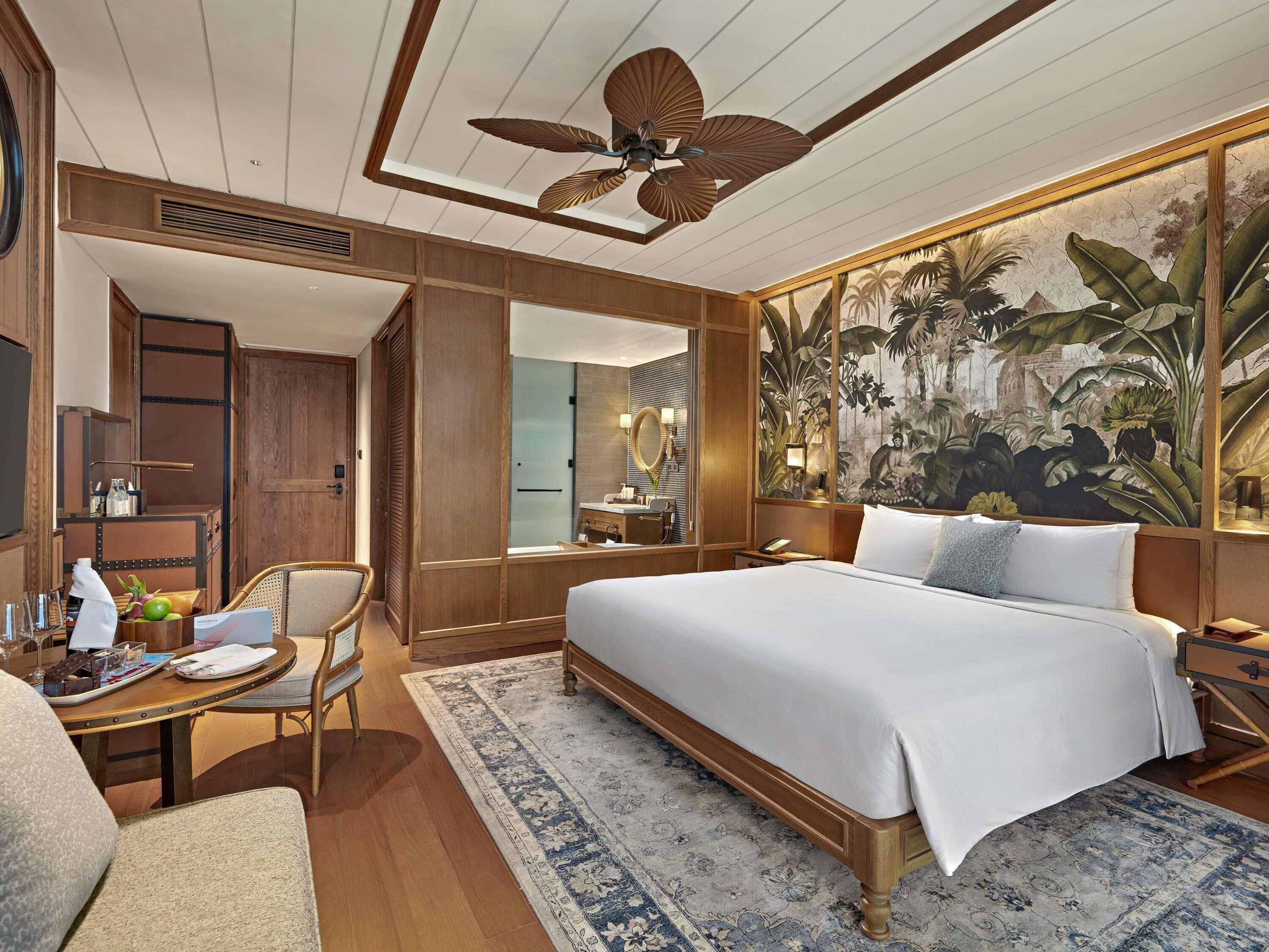 A hotel room with a bed, a desk with a chair, a large mirror, a ceiling fan, and a tropical mural.