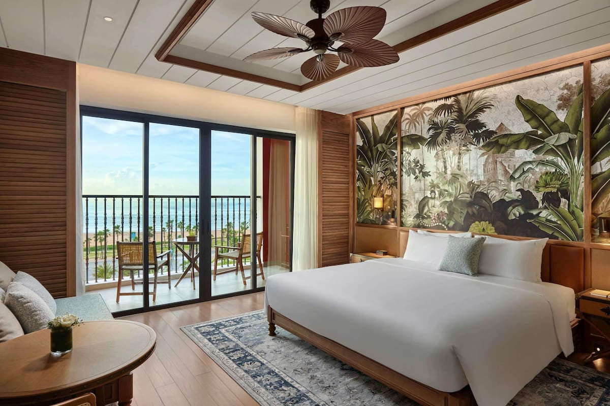 Habitación de hotel con una cama grande, un balcón con vista a la playa, un comedor con mesa y sillas, y un mural temático tropical.